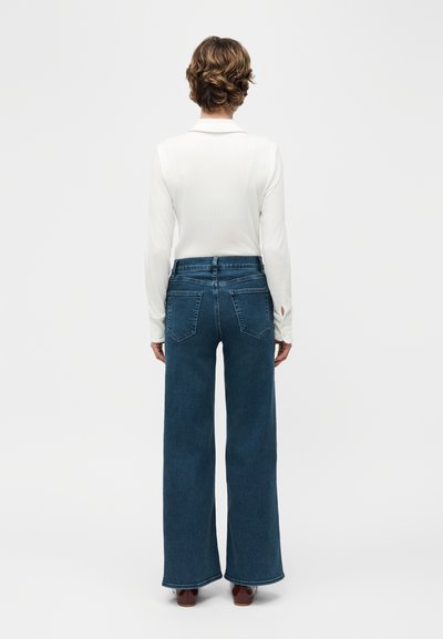 Άτομο με κοντά σγουρά μαλλιά που φοράει λευκό πουκάμισο με μακριά μανίκια, φαρδιά μπλε τζιν παντελόνια και καφέ παπούτσια, όρθιο με την πλάτη γυρισμένη.