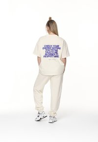 Krémové oversized tričko s fialovým textem na zadní straně. V kombinaci s odpovídajícími krémovými tepláky a bílými sportovními botami.