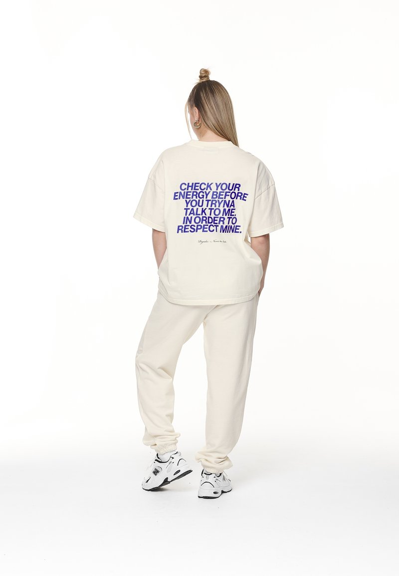 Krémové oversized tričko s fialovým textem na zadní straně. V kombinaci s odpovídajícími krémovými tepláky a bílými sportovními botami.