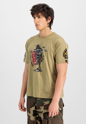 Jonge man met donker haar, die een olijfgroen T-shirt draagt met een zwart duivelmotief en camouflage cargobroek, staand tegen een witte achtergrond.