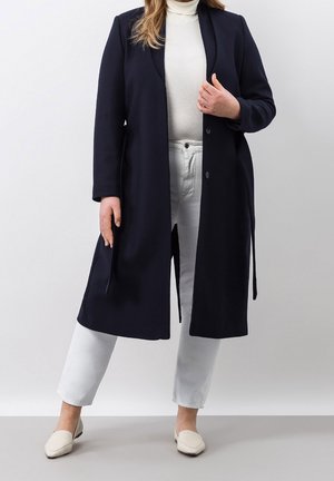 Mujer vistiendo un abrigo largo azul marino oscuro sobre un suéter de cuello alto blanco, pantalones blancos y zapatos blancos sin cordones, de pie frente a un fondo liso.