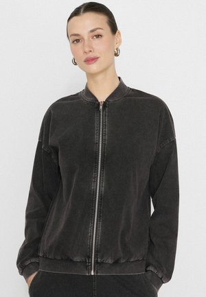 Młoda kobieta w ciemnej kurtce typu bomber na zamek i srebrnych kolczykach kołach, stojąca na jasnym, jednolitym tle, z rękami w kieszeniach.