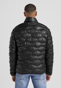 Schwarze Leder-Puffermantel mit gestepptem Textil, hohem Kragen und Logo-Patch auf dem Rücken. Kombiniert mit hellblauen Jeans.