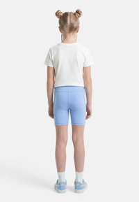 Παιδί με ξανθά μαλλιά σε διπλούς κότσους, φορώντας λευκό μπλουζάκι, γαλάζια σορτς, λευκές κάλτσες και μπλε αθλητικά παπούτσια, στέκεται γυρισμένο προς τα πίσω.