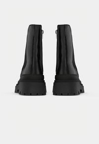 Paire de bottines noires à semelle épaisse, fermeture éclair au dos et texture cuir lisse, vues de dos sur fond blanc.