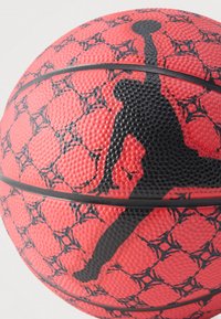 Közeli kép egy piros kosárlabdáról, fekete repedésmintával, valamint a labdával ugró kosárlabdázó fekete sziluettjéről.