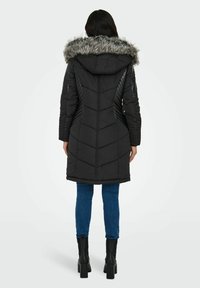 Manteau matelassé noir avec capuche bordée de fourrure, design matelassé et accents côtelés verticaux sur les épaules. Associé à un jean bleu et des bottes à talons noires.