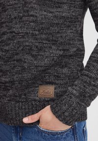 Strickenes Sweatshirt in Dunkelgrau- und Schwarztönen mit gerippten Bündchen und Saum, versehen mit einem braunen Lederetikett mit "Solid"-Markenlogo.