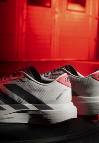 Paar weiße und schwarze Adidas Adizero Laufschuhe mit roten Akzenten, auf einer dunklen Oberfläche vor einem rot beleuchteten Hintergrund platziert.