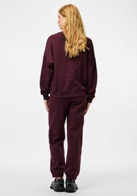 Oprijeta športna oblačila vinsko rdeče barve z ohlapnim krojem. Zgornji del ima spuščena ramena in rebraste manšete; hlače imajo elastike na spodnjem robu in stranske žepe.