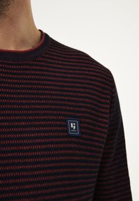 Suéter de punto a rayas en navy y rojo con un cuello redondo y un pequeño parche de marca azul en el pecho. Textura suave, diseño casual.