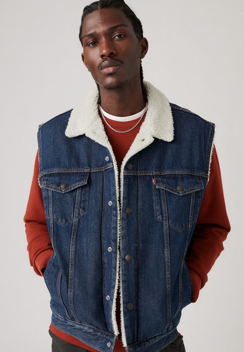 Denim vest med en lys fleecekrage, med knapper foran, to brystlommer, og en mørkeblå vask, lagt over en rød hettegenser.