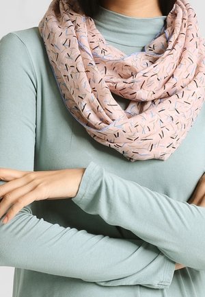 Personne portant une chemise à manches longues vert clair et une écharpe infinity rose à motifs, les bras croisés.