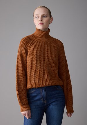 Chunky knit turtleneck sweater in burnt orange, featuring ribbed cuffs and a relaxed fit, paired with dark blue jeans.