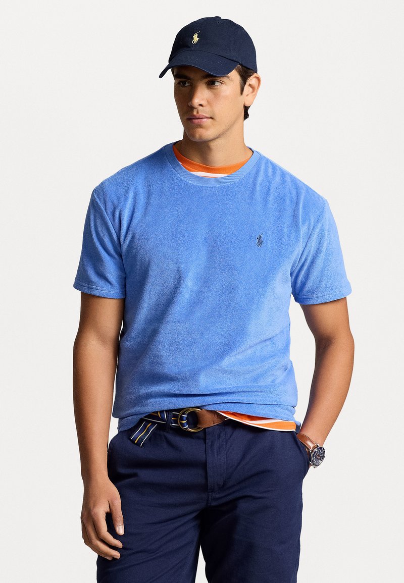 Tricou din bumbac frotir de culoare albastră, cu mâneci scurte, gât rotund și un mic logo brodat. Asortat cu pantaloni închiși la culoare și o șapcă bleumarin.