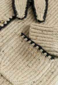 Wollcardigan mit einem strukturierten beigen Körper, schwarzem Saum und einer kleinen Tasche. Verfügt über einen Knopfverschluss und ein kontrastierendes, gemustertes Detail.