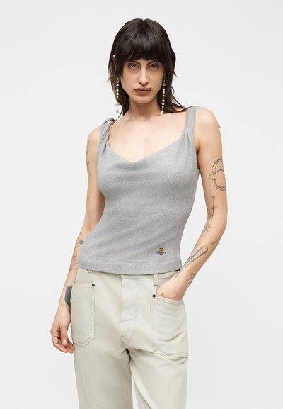 Γυναίκα με σκούρα μαλλιά μέχρι τους ώμους, που φοράει γκρι αμάνικη μπλούζα και ανοιχτό μπεζ παντελόνι, δείχνοντας τα τατουάζ στο χέρι και μακριά μαργαριταρένια σκουλαρίκια.