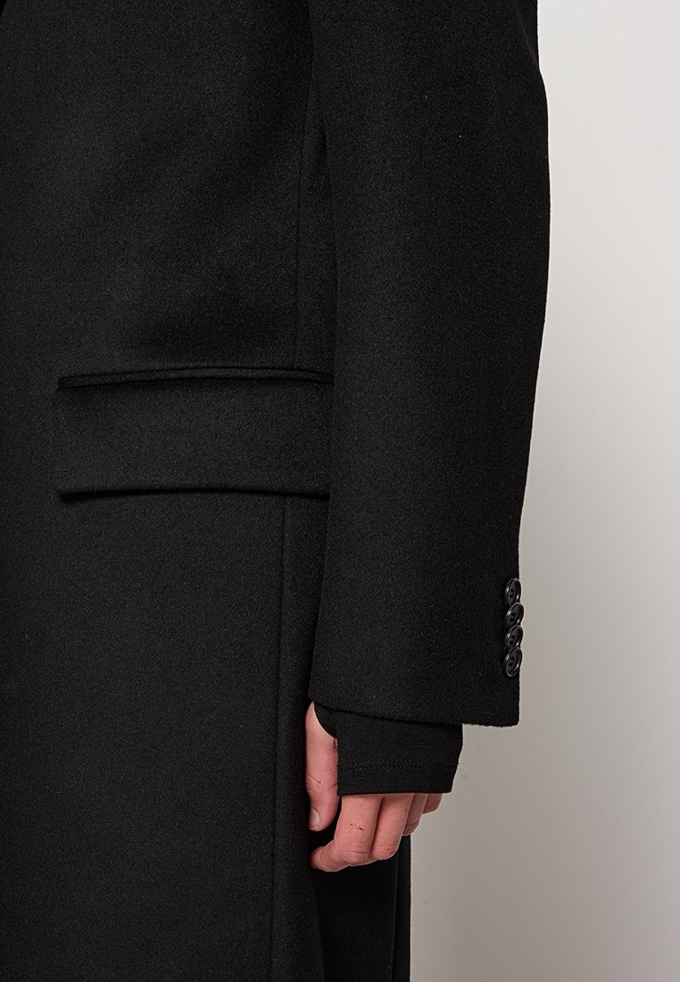Manteau en laine noire avec un design ajusté, poches latérales et détails de poignets à boutons sur les manches. Texture lisse, forme structurée.