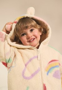 Hoodie en polaire beige avec des motifs abstraits colorés, y compris un arc-en-ciel. Présente des oreilles d'ours sur la capuche et une fermeture éclair à l'avant.