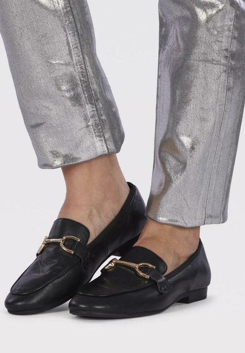 Des mocassins noirs en cuir présentent un détail en métal doré en forme de mors de cheval. Portés avec un pantalon argenté brillant arrivant aux chevilles, mettant en valeur des semelles plates et une texture lisse.