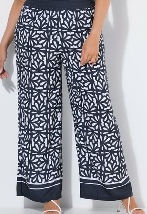 Femme portant un pantalon large imprimé noir et blanc avec une taille élastique et des sandales blanches à talons hauts et à brides.