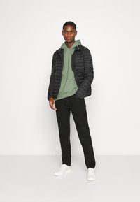 Black quilted jacket, green hoodie, black pants, and white sneakers. Model stands with hands relaxed, on a plain background.