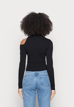 Mujer con cabello rizado que lleva una blusa negra de manga larga con un corte en un hombro y jeans azules de talle alto, vista desde atrás.