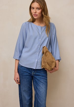 Junge Frau in blau-weiß gestreifter Bluse mit Knöpfen und blauen Jeans hält eine weiche braune Wildledertasche vor einem neutralen Hintergrund.