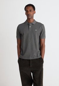 Graues Poloshirt mit zwei Knöpfen am Kragen, kurzen Ärmeln und einem dezenten Logo auf der Brust, kombiniert mit dunklen Cordhosen.