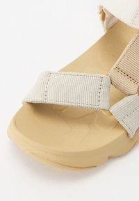Beige sandal featuring textured rubber sole, adjustable fabric straps in a light tone, and a padded design for comfort and support.