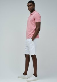 Polo de color rosa claro, pantalones cortos blancos y zapatillas blancas. El polo tiene cuello y mangas cortas. El conjunto tiene un aspecto casual y moderno.