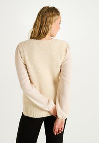 Pull en tricot beige avec une finition texturée, ourlet côtelé et encolure ronde, superposé sur une blouse légère en tissu transparent à manches longues.
