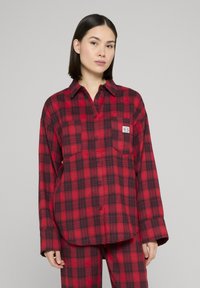 Chemise en flanelle à carreaux rouge et noir avec manches longues, fermeture à boutons, deux poches avant et un petit patch logo sur le côté gauche.