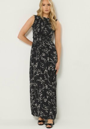 Frau trägt ein ärmelloses schwarzes Maxikleid mit weißen Blumenmustern, steht mit einer Hand in der Hüfte vor einem einfarbigen Hintergrund.