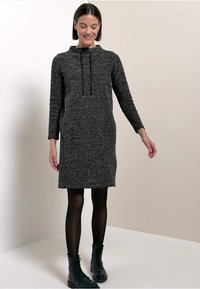 Šedé pletené šaty s kapucí, dlouhými rukávy, detaily na stahování a volným střihem, v kombinaci s černými šněrovacími botami a průhlednými černými punčochami.