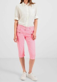 Camisa blanca ligera de botones con mangas arremangadas, combinada con pantalones cortos rosas que tienen cremalleras en los bolsillos frontales y zapatillas blancas.