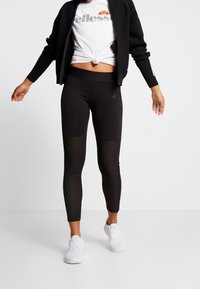 Sorte atletiske leggings med netpaneler, parret med en hvid t-shirt og en sort zip-up jakke, båret med hvide sneakers.