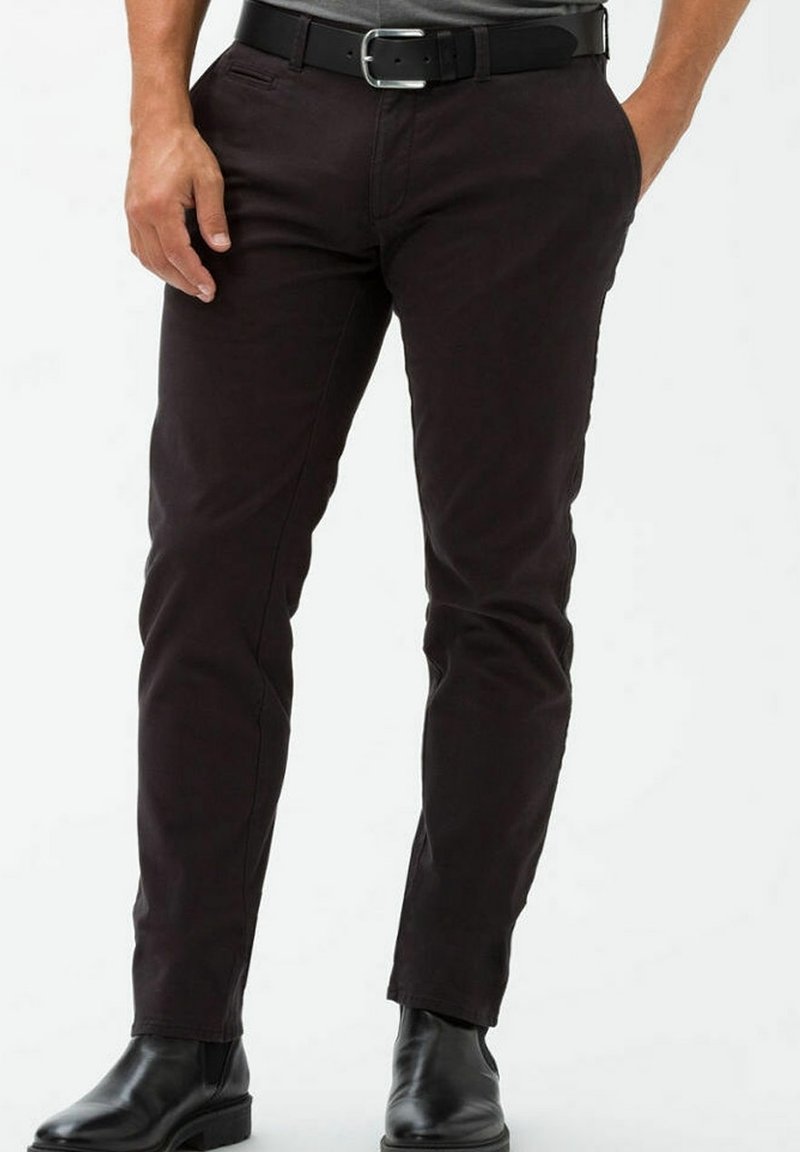 Homme portant un pantalon noir ajusté, une ceinture en cuir noire et des bottes en cuir noires, debout devant un fond uni.