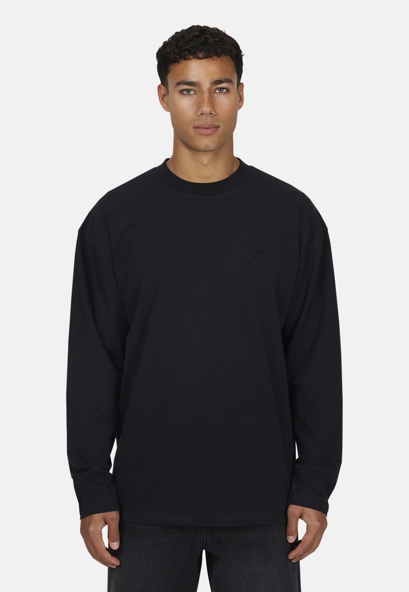 Jonge man met kort krullend haar in een effen zwart longsleeve-shirt en donkere broek, staande tegen een lichtgrijze achtergrond.