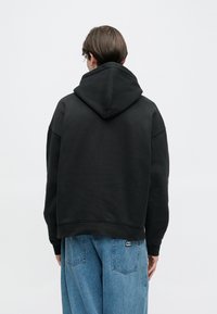 Fekete kapucnis pulóver nagy kapucnival és laza szabással, bordázott szegéllyel és mandzsettákkal. Az anyag puha és texturált hatású.