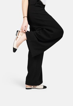 Vrouw die zwarte broek met wijde pijpen draagt en beige en zwarte slingback platte schoenen met een kleine gesp aan het enkelbandje verstelt.