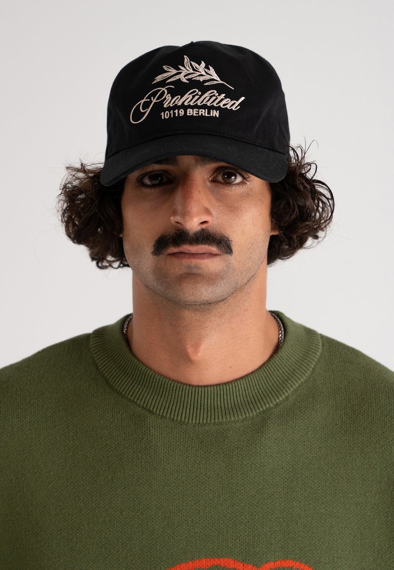 Svart baseballcap med hvit brodert "Prohibited 10119 Berlin" og bladdesign. Modellen har på seg en grønn strikkegenser med oransje mønster.