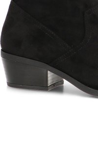 Botín negro de ante hasta el tobillo con un tacón geométrico apilado. Textura suave y sutil detalle de costura en la parte superior.