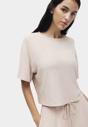 Ung kvinde iført lys beige ribstrikket kortærmet top og matchende bukser med snoretræk, stående foran en ensfarvet hvid baggrund.