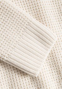 Dettaglio di un maglione lavorato a maglia di colore crema, caratterizzato da un corpo texturizzato con un motivo a nido d'ape e polsini a costine per una maggiore struttura.