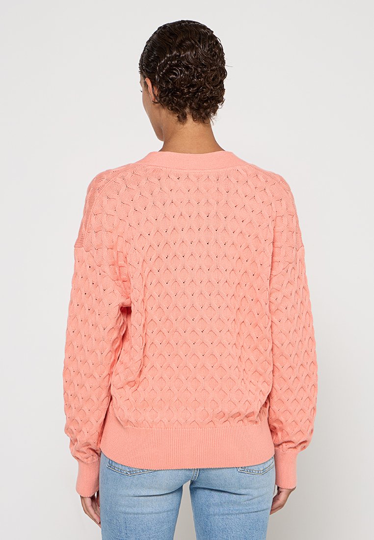 Różowy sweter dzianinowy z wzorem w romby, z bufkami i ściągaczem u dołu. Nosi się go na jasnoniebieskich dżinsach. Miękka faktura i luźny krój.