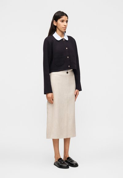 Γυναίκα στέκεται φορώντας μαύρο σακάκι με κουμπιά πάνω από ένα πουκάμισο με γιακά, μ πεζ μίντι φούστα και μαύρα μοκασίνια χωρίς κορδόνια, μπροστά σε ένα απλό φωτεινό φόντο.