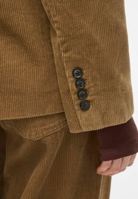 Manteau en velours côtelé marron avec des manches comportant quatre boutons noirs, présentant une texture côtelée et un tissu bordeaux contrastant en dessous.