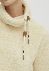 Cream-colored fleece pullover with a high collar and drawstrings. Textured, soft fabric with brown wooden accents on the drawstrings.
