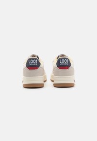 Lacoste L001 sneaker bézs és szürke velúrral, piros és tengerészkék részletekkel, mintás gumitalppal és dombornyomott logóval a sarkán.
